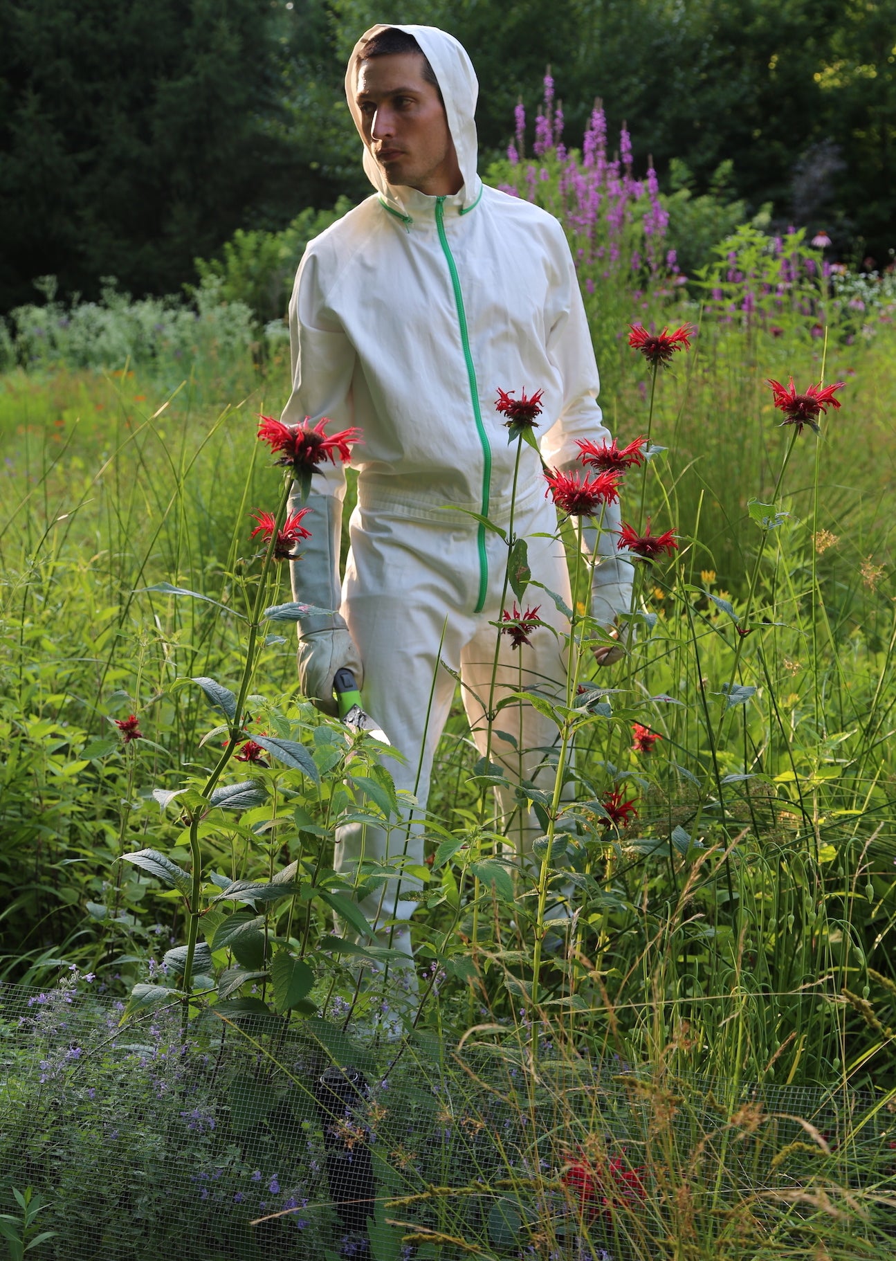 Man standing in tall grass wearing TheTickSuit holding a pair of gardening scissors.